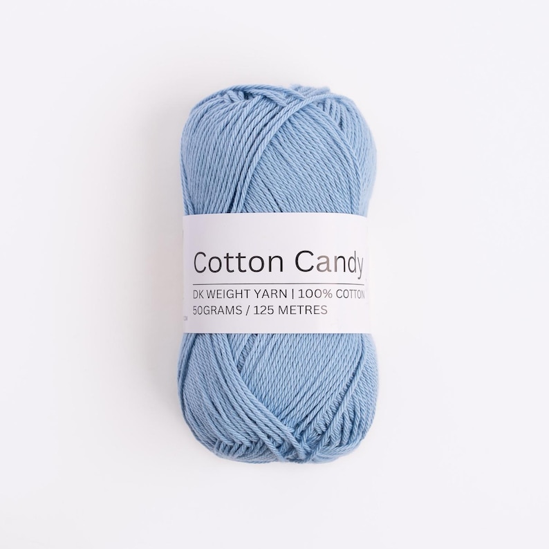 May include: A skein of light blue DK weight yarn wrapped in a white paper label that reads Cotton Candy, DK WEIGHT YARN, 100% COTTON, 50 GRAMS, 125 METRES. The yarn has a smooth, twisted texture and is presented in a compact, oval-shaped bundle.