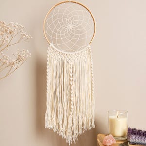 May include: A cream-colored dreamcatcher with a woven web design and long fringe hangs against a neutral wall. The dreamcatcher has a wooden hoop and macrame detailing. A lit candle and crystals are in the background.