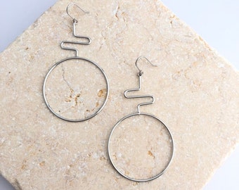 Puede incluir: Un par de pendientes de aro geométricos de plata con un diseño de círculo y línea curva. Los pendientes se muestran sobre una superficie de piedra beige claro.