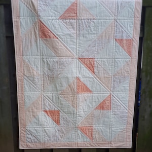 Może przedstawiać: Różowa i biała kołdra z geometrycznym wzorem trójkątów. Kołdra jest zawieszona na drewnianym płocie.