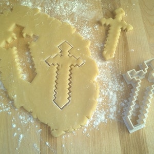 May include: A light brown, uncooked dough with a sword-shaped cutout and two sword-shaped cookies. A white plastic sword-shaped cookie cutter is also present. The scene is set on a wooden cutting board with scattered flour.