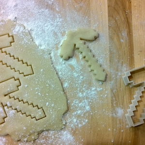 May include: A wooden cutting board with cookie dough, flour, and pickaxe-shaped cookie cutters. Two pickaxe cookies are cut out of the dough, one is baked, and one is a cookie cutter.