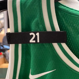 May include: Green basketball jersey with white and green striped trim. The number "21" is displayed in white on a black nameplate. A white Nike swoosh logo is visible on the jersey.