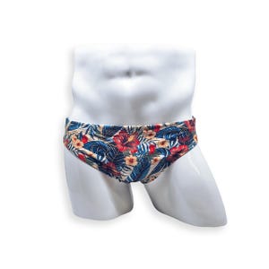 Op de afbeelding: Zwemslip met een tropisch bloemenpatroon. De slip heeft een beige achtergrond met rode hibiscusbloemen, blauwe bladeren en ander tropisch gebladerte. De badkleding wordt getoond op een witte mannequin.