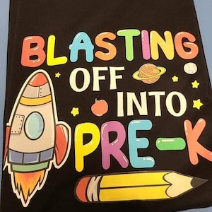 May include: A black t-shirt with a colorful graphic design featuring a rocket, a pencil, and the text "Blasting Off Into Pre-K".