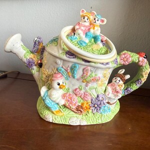 Könnte beinhalten: Eine dekorative Gießkanne aus Keramik mit einem skurrilen Gartenthema. Die Kanne ist mit bunten Blumen, Schmetterlingen und charmanten Tierfiguren verziert, darunter eine Ente, ein Hase und zwei Mäuse. Der Griff und der Ausguss sind mit floralen Designs versehen.