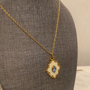 May include: A gold-toned necklace with a pendant featuring a white, pearl-like border and a central, oval-shaped blue stone. The pendant has a decorative, ornate design. The necklace is displayed on a gray surface.