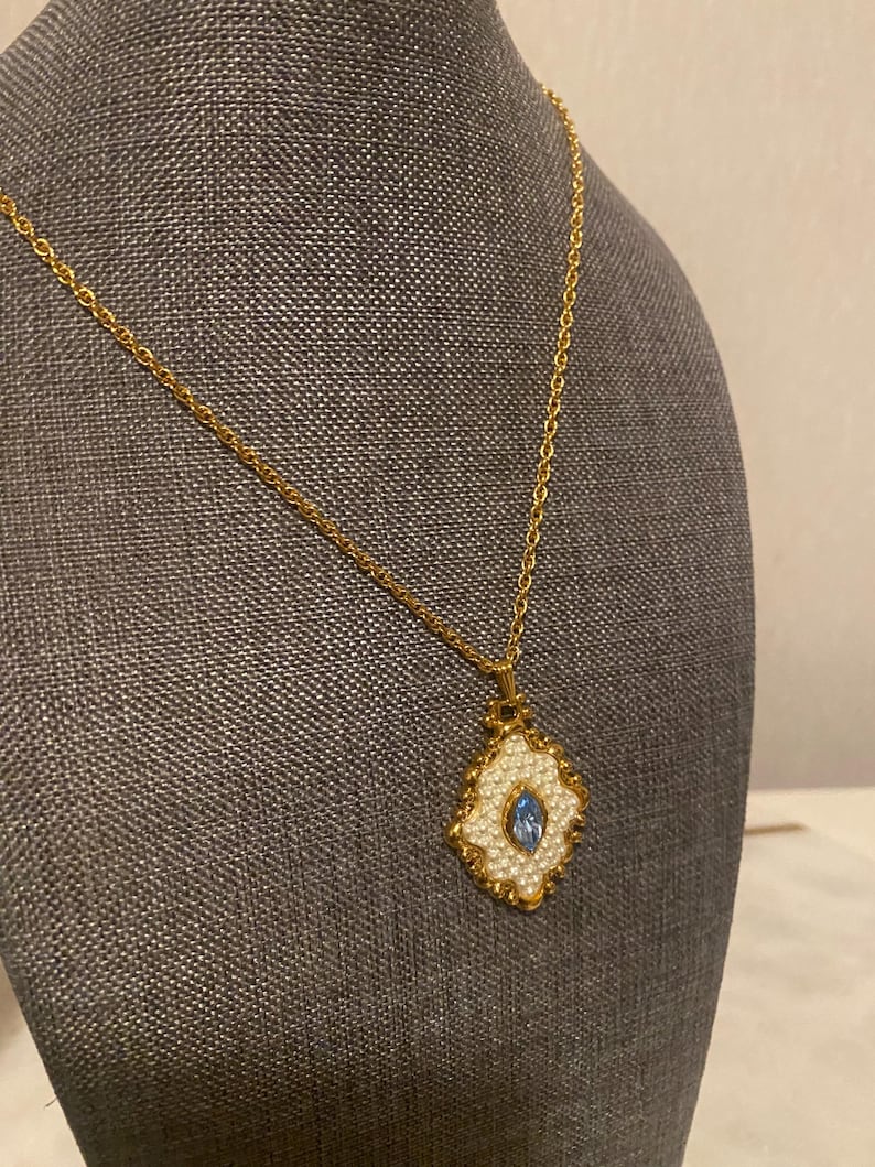 May include: A gold-toned necklace with a pendant featuring a white, pearl-like border and a central, oval-shaped blue stone. The pendant has a decorative, ornate design. The necklace is displayed on a gray surface.