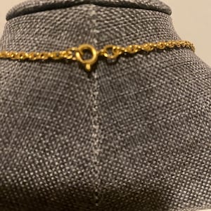 May include: Close-up of a gold-colored necklace with a delicate chain and a circular clasp. The necklace is displayed on a gray mannequin. The clasp is open, showing the details of the design.
