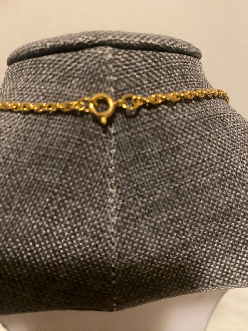 May include: Close-up of a gold-colored necklace with a delicate chain and a circular clasp. The necklace is displayed on a gray mannequin. The clasp is open, showing the details of the design.