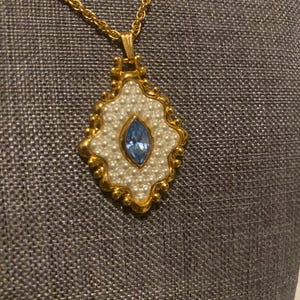 May include: Gold-toned necklace with a pendant featuring a central blue gemstone surrounded by faux pearls. The pendant has a gold-colored ornate frame. The chain is also gold-toned.