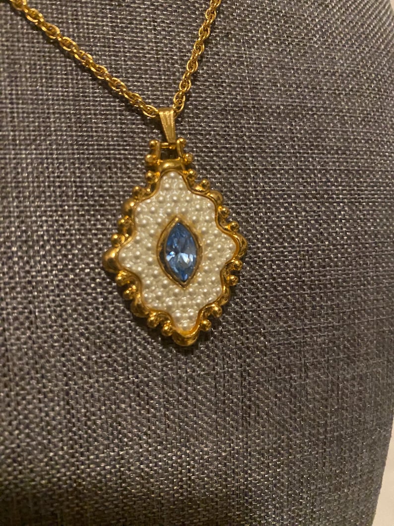 May include: Gold-toned necklace with a pendant featuring a central blue gemstone surrounded by faux pearls. The pendant has a gold-colored ornate frame. The chain is also gold-toned.