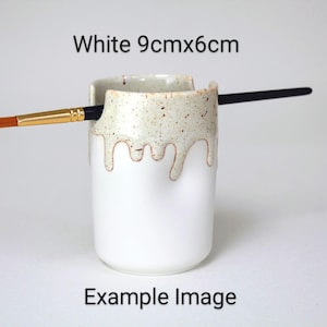 A White porcelain pot with a cream speckled glaze dripping down the top third. Two cut out grooves on opposite sides of the rim, with a paintbrush resting inside. Halfway down the pot on opposite sides are a thumb and finger indent for grip.