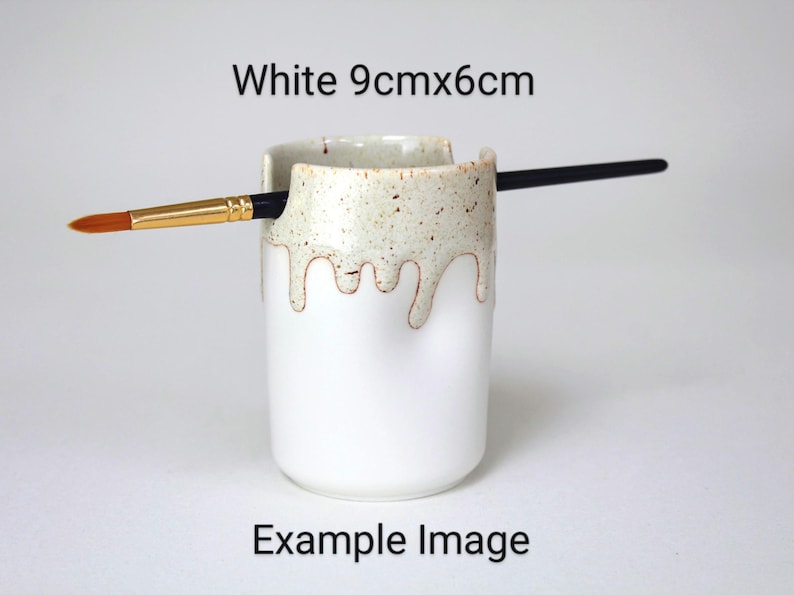 A White porcelain pot with a cream speckled glaze dripping down the top third. Two cut out grooves on opposite sides of the rim, with a paintbrush resting inside. Halfway down the pot on opposite sides are a thumb and finger indent for grip.
