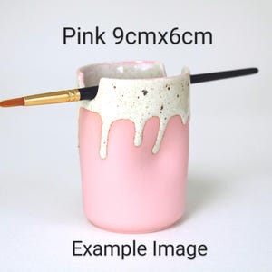 A Pink porcelain pot with a cream speckled glaze dripping down the top third. Two cut out grooves on opposite sides of the rim, with a paintbrush resting inside. Halfway down the pot on opposite sides are a thumb and finger indent for grip.