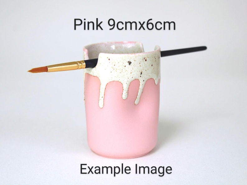 A Pink porcelain pot with a cream speckled glaze dripping down the top third. Two cut out grooves on opposite sides of the rim, with a paintbrush resting inside. Halfway down the pot on opposite sides are a thumb and finger indent for grip.