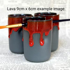 May include: Three gray ceramic mugs with red glaze dripping down the sides. The mugs are 9 cm by 6 cm and are arranged in a row. A paintbrush is resting on top of the mugs.
