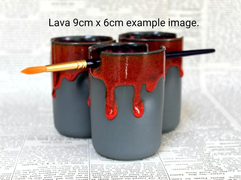May include: Three gray ceramic mugs with red glaze dripping down the sides. The mugs are 9 cm by 6 cm and are arranged in a row. A paintbrush is resting on top of the mugs.
