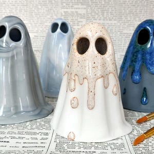 May include: A selection of ceramic ghost ornaments in shades of grey, blue, and white. The ghosts feature large, dark eye holes and a draped, flowing form. Paintbrushes are present in the foreground.