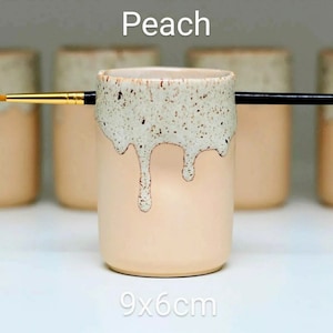 A Peach porcelain pot with a cream speckled glaze dripping down the top third. Two cut out grooves on opposite sides of the rim, with a paintbrush resting inside. Halfway down the pot on opposite sides are a thumb and finger indent for grip.