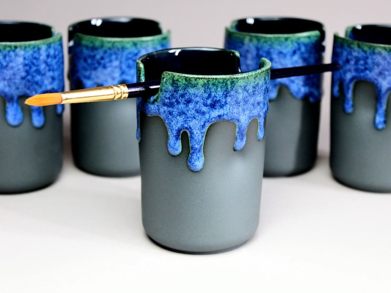 May include: Five gray ceramic mugs with a blue and green glaze. The glaze has a drip effect. A paintbrush is resting on the rim of one of the mugs.