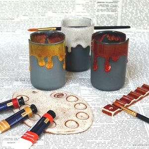 May include: Three ceramic cups with dripping glaze in yellow, red, and white. The cups are on a newspaper background with paint tubes and a paint palette.
