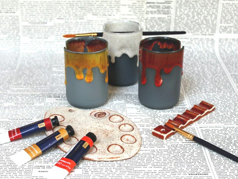 May include: Three ceramic cups with dripping glaze in yellow, red, and white. The cups are on a newspaper background with paint tubes and a paint palette.