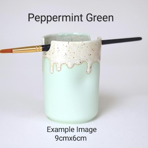 A Peppermint Green porcelain pot with a cream speckled glaze dripping down the top third. Two cut out grooves on opposite sides of the rim, with a paintbrush resting inside. Halfway down the pot on opposite sides are a thumb and finger indent.