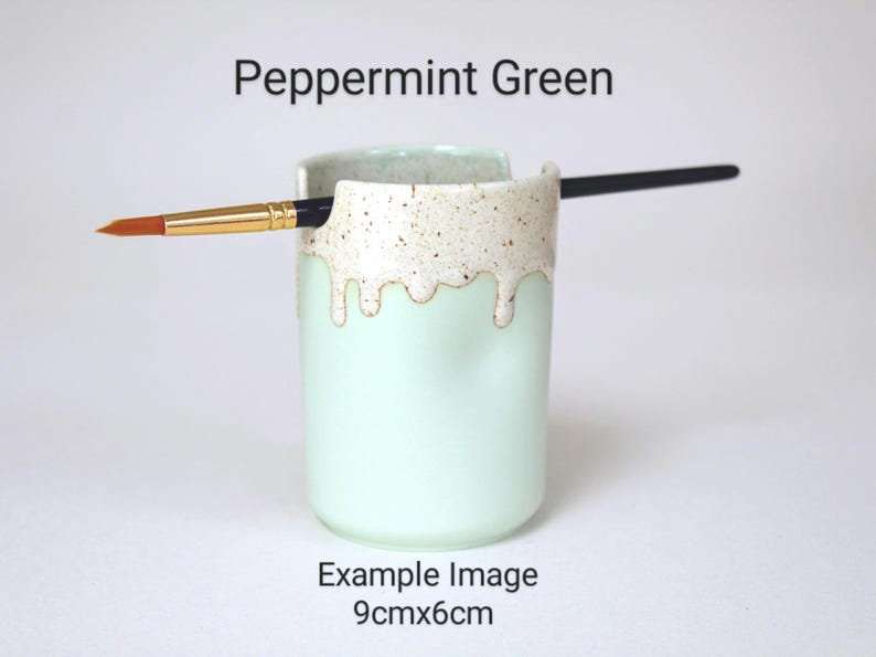 A Peppermint Green porcelain pot with a cream speckled glaze dripping down the top third. Two cut out grooves on opposite sides of the rim, with a paintbrush resting inside. Halfway down the pot on opposite sides are a thumb and finger indent.