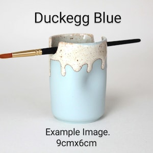 A Duckegg Blue porcelain pot with a cream speckled glaze dripping down the top third. Two cut out grooves on opposite sides of the rim, with a paintbrush resting inside. Halfway down the pot on opposite sides are a thumb and finger indent for grip.