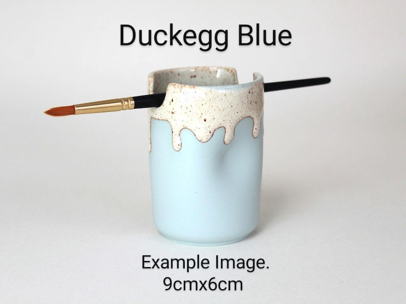 A Duckegg Blue porcelain pot with a cream speckled glaze dripping down the top third. Two cut out grooves on opposite sides of the rim, with a paintbrush resting inside. Halfway down the pot on opposite sides are a thumb and finger indent for grip.