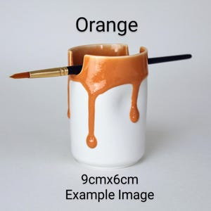 May include: Orange ceramic brush holder with a drip glaze design. The holder is 9cm x 6cm and is shown with a paintbrush.