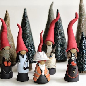 May include: A collection of ceramic gnomes with red pointed hats and various outfits, standing in front of textured ceramic Christmas trees. The gnomes have long, curved noses and detailed beards. Some gnomes are wearing aprons, and one has a construction hat.