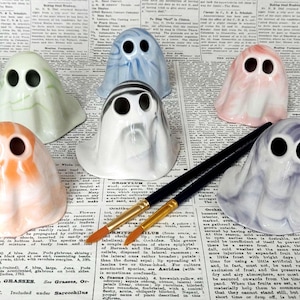 May include: A collection of ceramic ghost figurines in various colors, including orange, blue, pink, and white, with large black eye holes. Two paintbrushes with black handles and gold ferrules are placed near the ghosts. The ghosts are on a newspaper background.