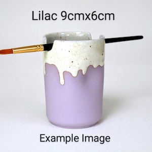 A Lilac porcelain pot with a cream speckled glaze dripping down the top third. Two cut out grooves on opposite sides of the rim, with a paintbrush resting inside. Halfway down the pot on opposite sides are a thumb and finger indent for grip.