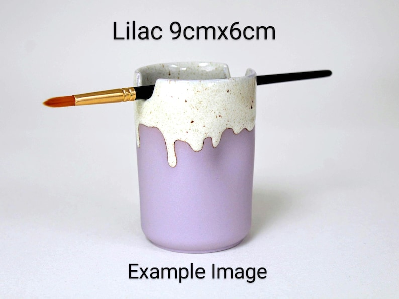 A Lilac porcelain pot with a cream speckled glaze dripping down the top third. Two cut out grooves on opposite sides of the rim, with a paintbrush resting inside. Halfway down the pot on opposite sides are a thumb and finger indent for grip.
