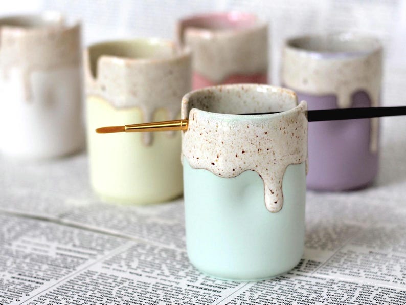 Coloured porcelain pots with a cream speckled glaze dripping down the top third. Two cut out grooves on opposite sides of the rim, with a paintbrush resting inside. Halfway down the pot on opposite sides are a thumb and finger indent.