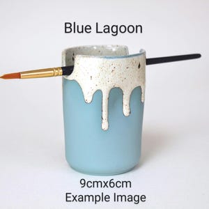 May include: Blue ceramic brush holder with a white drip glaze. The holder is 9 cm by 6 cm and has a brush resting in it.