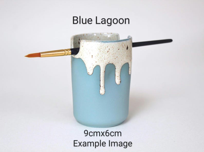 May include: Blue ceramic brush holder with a white drip glaze. The holder is 9 cm by 6 cm and has a brush resting in it.