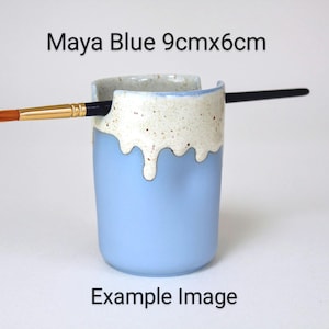 A Maya Blue porcelain pot with a cream speckled glaze dripping down the top third. Two cut out grooves on opposite sides of the rim, with a paintbrush resting inside. Halfway down the pot on opposite sides are a thumb and finger indent for grip.