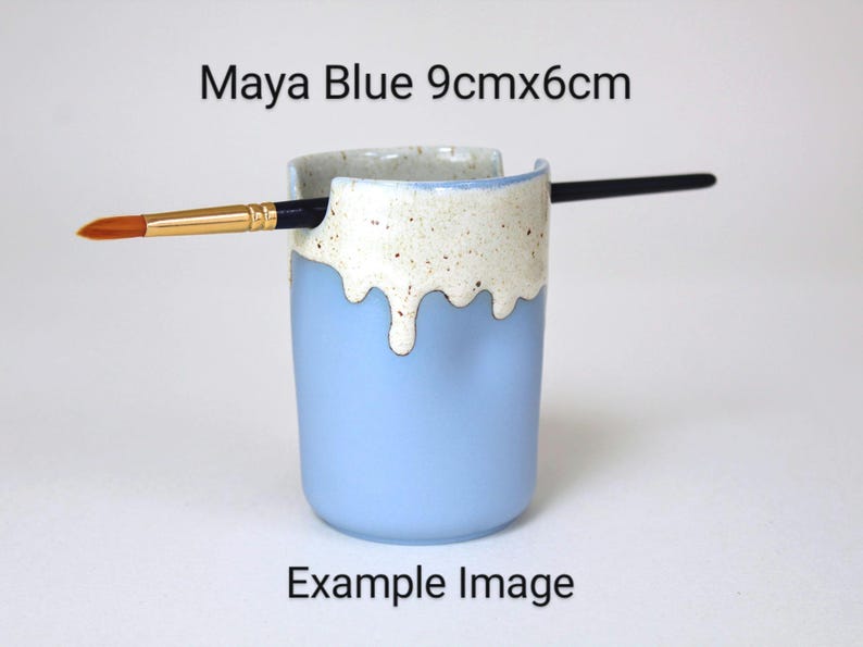 A Maya Blue porcelain pot with a cream speckled glaze dripping down the top third. Two cut out grooves on opposite sides of the rim, with a paintbrush resting inside. Halfway down the pot on opposite sides are a thumb and finger indent for grip.