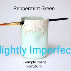 Könnte beinhalten: Ein minzgrüner Keramik-Pinselhalter mit einem gesprenkelten weißen Tropfglasur-Muster am oberen Rand. Ein Pinsel mit schwarzem Stiel liegt in der Einkerbung. Der Text lautet Peppermint Green, Slightly Imperfect und 9cmx6cm.