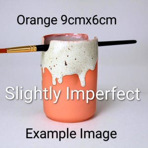 May include: A cylindrical orange ceramic brush holder measuring 9cm by 6cm, featuring a white speckled glaze dripping down from the top rim. A thin paintbrush with a gold ferrule rests horizontally across the notched top of the container.