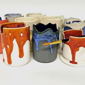 May include: A selection of ceramic paint holders in various colours, including orange, blue, and grey. Some holders have a dripped paint effect. A paintbrush rests in one of the holders, suggesting their use for art supplies.
