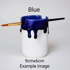 May include: A white ceramic paintbrush holder with a blue glaze dripping down the sides. The holder is 9 cm by 6 cm.