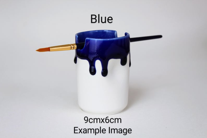 May include: A white ceramic paintbrush holder with a blue glaze dripping down the sides. The holder is 9 cm by 6 cm.