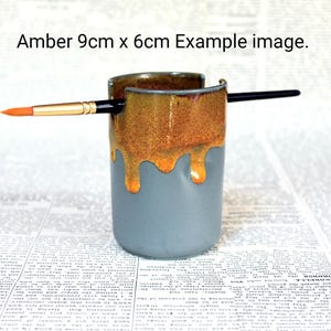 May include: A ceramic brush holder with a drip glaze in shades of amber and blue. The holder is 9 cm by 6 cm and has a brush resting on the top.