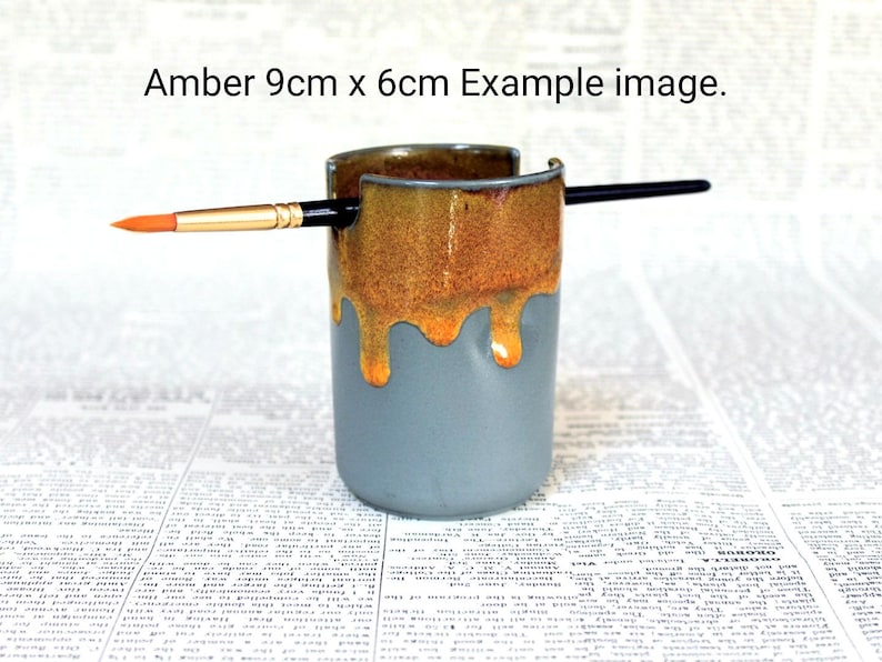 May include: A ceramic brush holder with a drip glaze in shades of amber and blue. The holder is 9 cm by 6 cm and has a brush resting on the top.