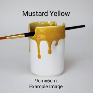 May include: A white ceramic brush holder with a mustard yellow glaze dripping down the sides. The holder is 9 cm by 6 cm and has a brush resting inside.