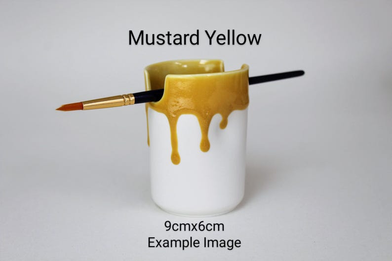 May include: A white ceramic brush holder with a mustard yellow glaze dripping down the sides. The holder is 9 cm by 6 cm and has a brush resting inside.